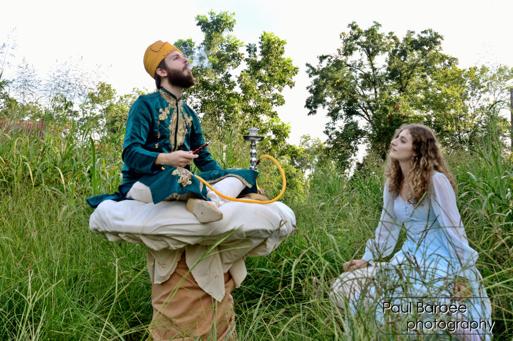 A man sitting on a mushroom blowing smoke while a woman looks at him. They are in a field